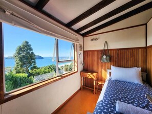 A bedroom with a bed, a desk, and a window with a view of the sea.