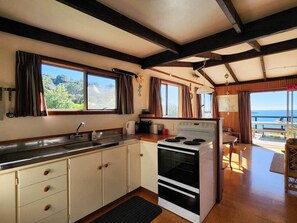A kitchen with a stove, sink, and cabinets, a dining area with a table and chairs, and a view of the sea through large windows. - Classic Kiwi Oceanview Bach (Hahei)