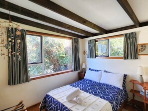 A bedroom with a bed, two windows, curtains, a bedside table, and a lamp. - Classic Kiwi Oceanview Bach (Hahei)