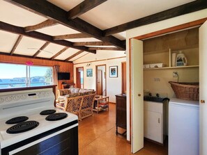 A kitchen with a stove, a refrigerator, and a dining area with a table and chairs. - Classic Kiwi Oceanview Bach (Hahei)