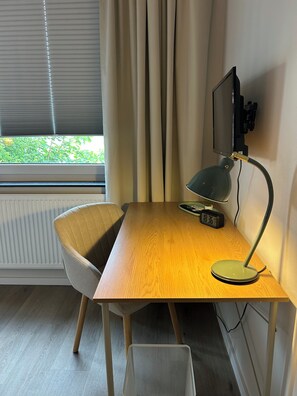 A room with a wooden desk, a chair, a desk lamp, a television, and a window with curtains.