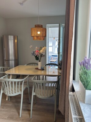 A modern kitchen with a stainless steel refrigerator, a dining table with chairs, a hanging light fixture, and a window with curtains.