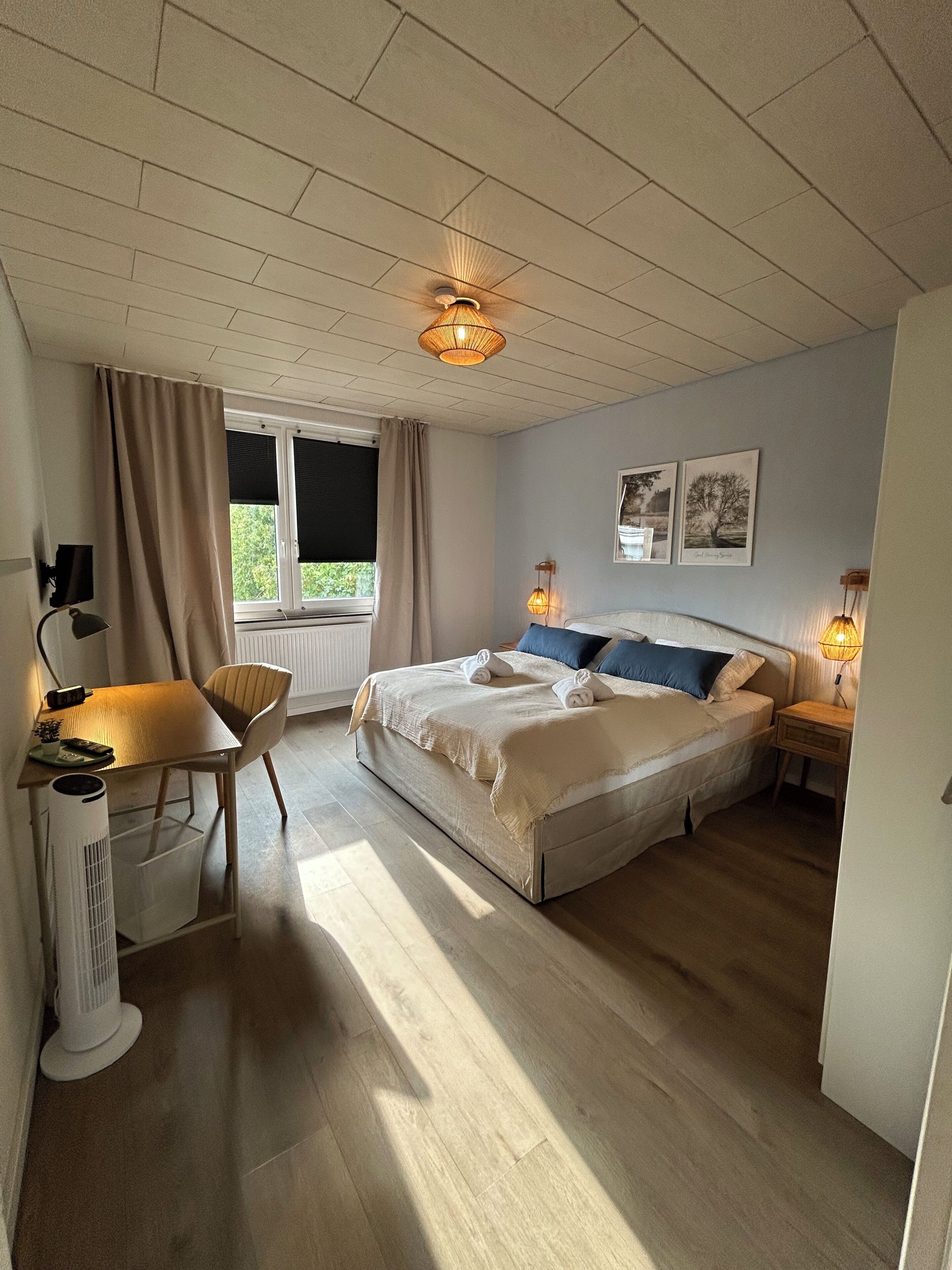 A bedroom with a bed, bedside tables, a desk, a chair, a window, and a ceiling fan.