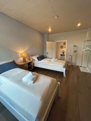 A hotel room with two beds, a bedside table with a lamp, a wardrobe, and a door leading to another room.