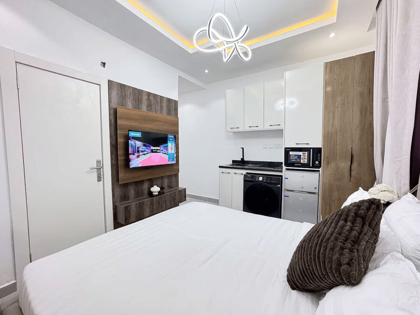 Une chambre à coucher moderne avec un grand lit, un téléviseur fixé au mur et une petite cuisine en arrière-plan.