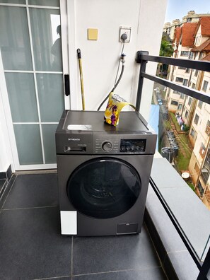 Un lave-linge compact et moderne, installé sur un balcon avec vue sur la ville, un petit sac jaune et une prise de courant.