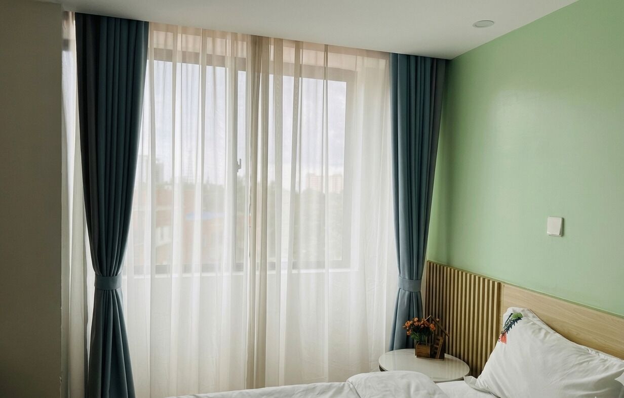 A hotel room with a bed, a bedside table with a vase of flowers, and sheer curtains.