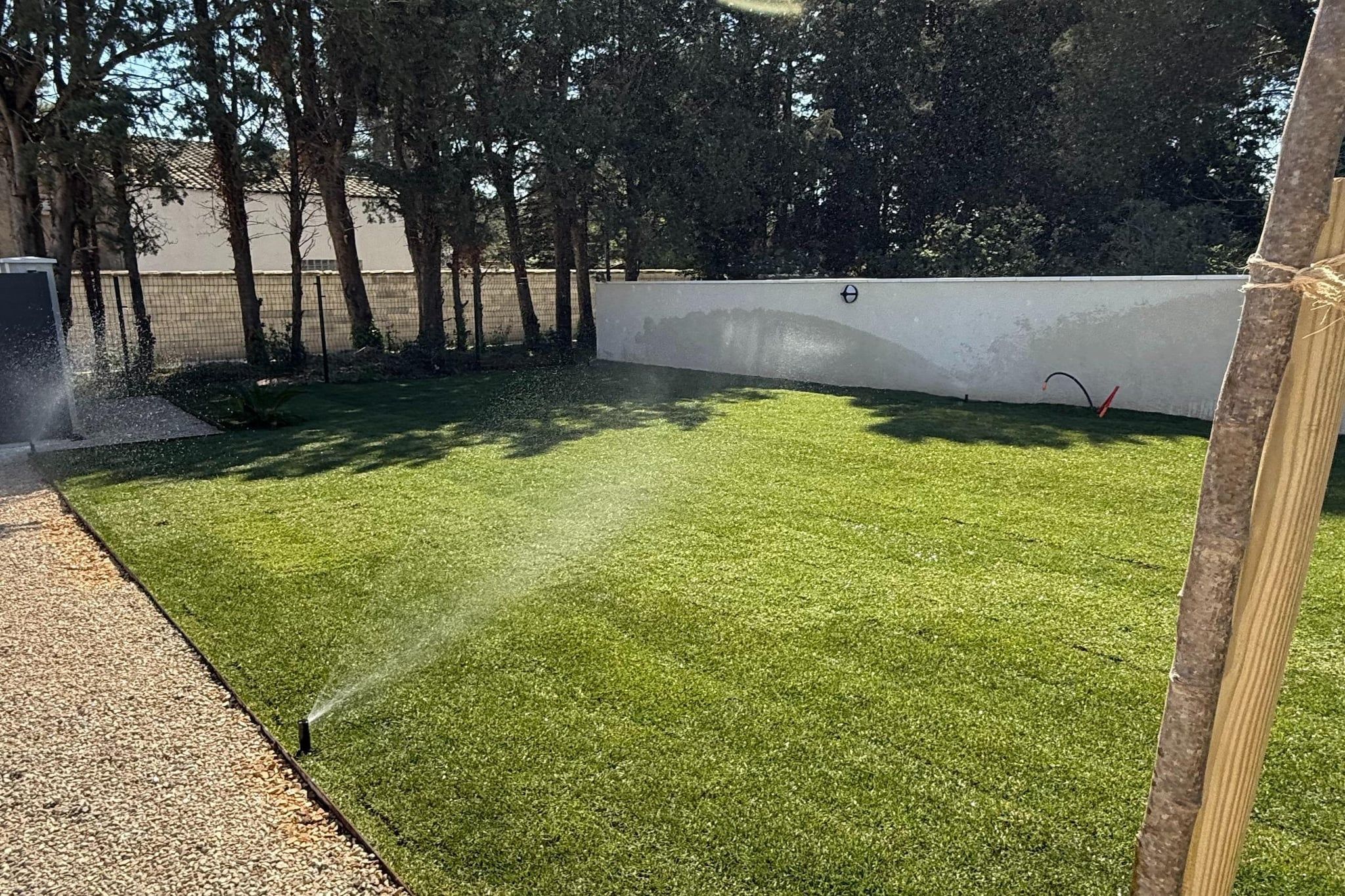A well-maintained artificial grass lawn with a sprinkler system, surrounded by a white wall and trees.