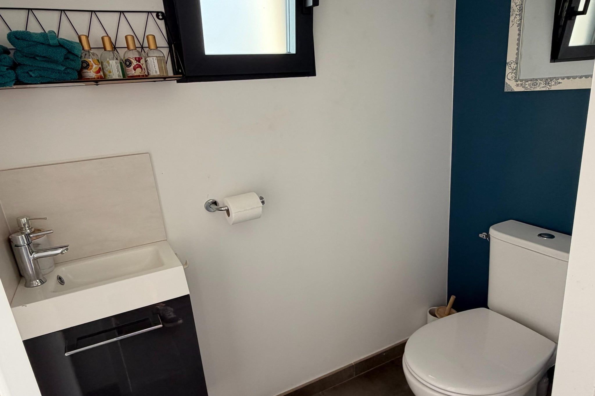 Une petite salle de bain avec un lavabo blanc, des toilettes et une petite fenêtre située au-dessus du lavabo.