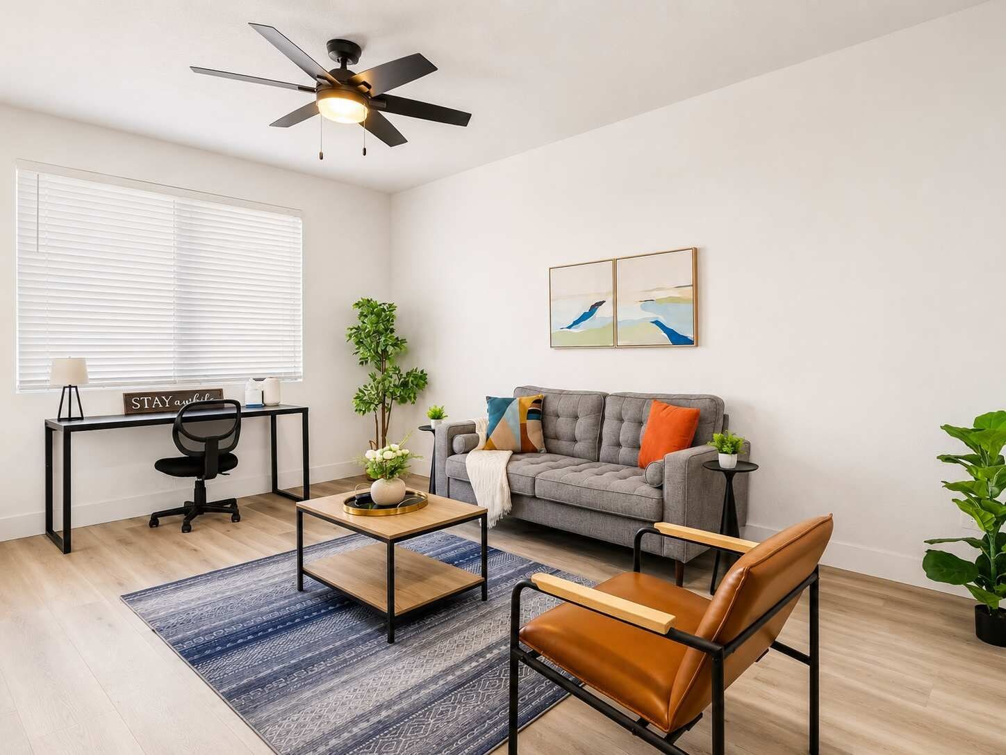 Ein modernes Wohnzimmer mit grauem Sofa, einem Holztisch, einem Ledersessel, einem Schreibtisch mit Stuhl, einem Deckenventilator und einem Fenster mit Jalousien.
