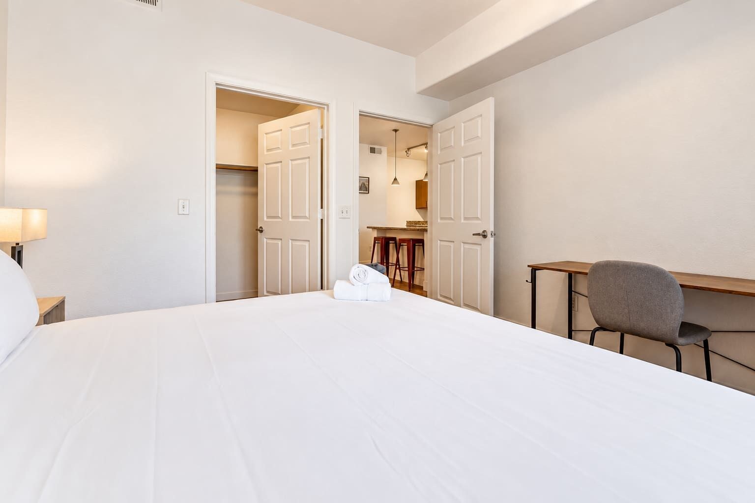 Une chambre d’hôtel avec un grand lit, un bureau et une chaise. Deux portes donnent sur une petite cuisine et une salle de bain.