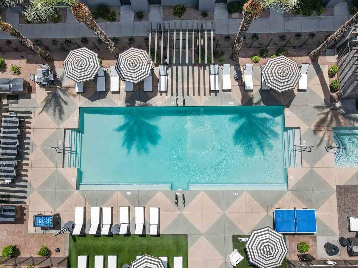 An aerial view of a swimming pool area with multiple lounge chairs and umbrellas, surrounded by palm trees and a tiled deck.