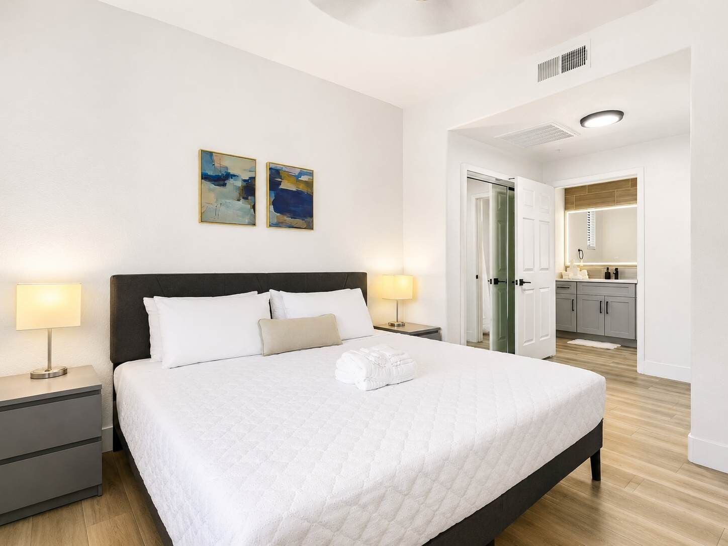 Habitación de hotel con una cama grande, dos lámparas de noche, una mesita de noche, un armario y un baño visible a través de una puerta abierta.