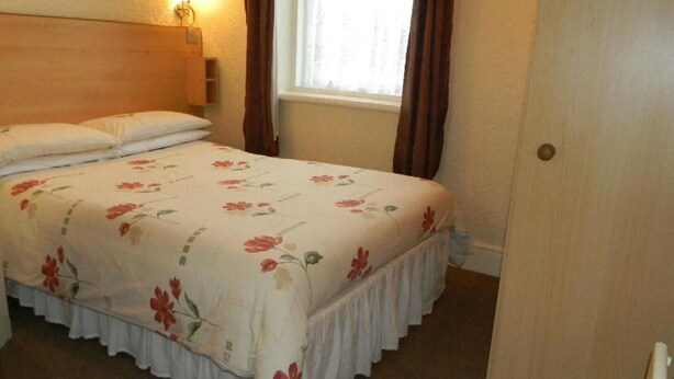 A hotel room with a bed, a nightstand, a wall-mounted lamp, and a window with curtains. - Standard Triple Room (Blackpool)