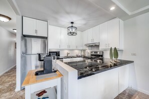 A modern kitchen with white cabinets, a stainless steel refrigerator, a black marble island, and a central island with a stove. - Remodeled 1 Bedroom Across from Kamaole Beach III–Kamaole Sands 10202 (Kihei)