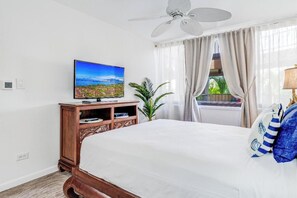 A bedroom with a bed, a television on a wooden stand, a ceiling fan, and a window with curtains. - Remodeled 1 Bedroom Across from Kamaole Beach III–Kamaole Sands 10202 (Kihei)