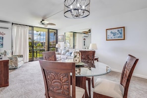 A spacious living room with a glass dining table, wooden chairs, a chandelier, and a view of the outdoors. - Remodeled 1 Bedroom Across from Kamaole Beach III–Kamaole Sands 10202 (Kihei)