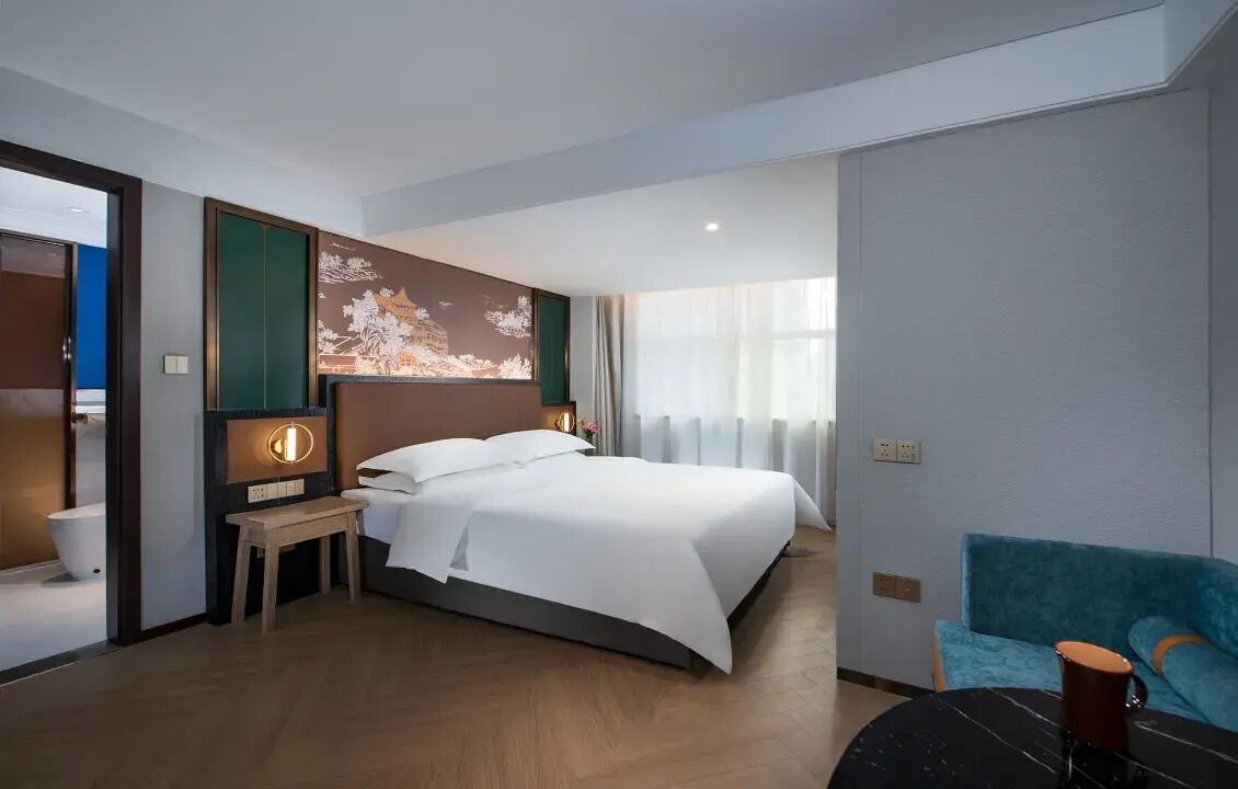 A hotel room with a bed, bedside table, lamp, and a view of the outside.