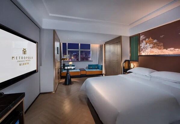 A hotel room with a bed, a desk, a chair, a sofa, a TV, and a window with a city view.