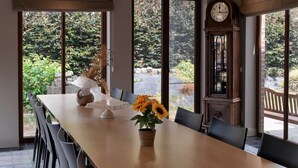 Een eethoek met een lange tafel, stoelen, een vaas met zonnebloemen, een klok en uitzicht op groen.