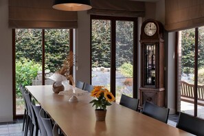 A dining area with a long table, chairs, a vase with sunflowers, a clock, and a view of greenery outside. - Apartment 3 – Enjoy a cozy stay at the Alfonsinehoeve (Horpmaal)