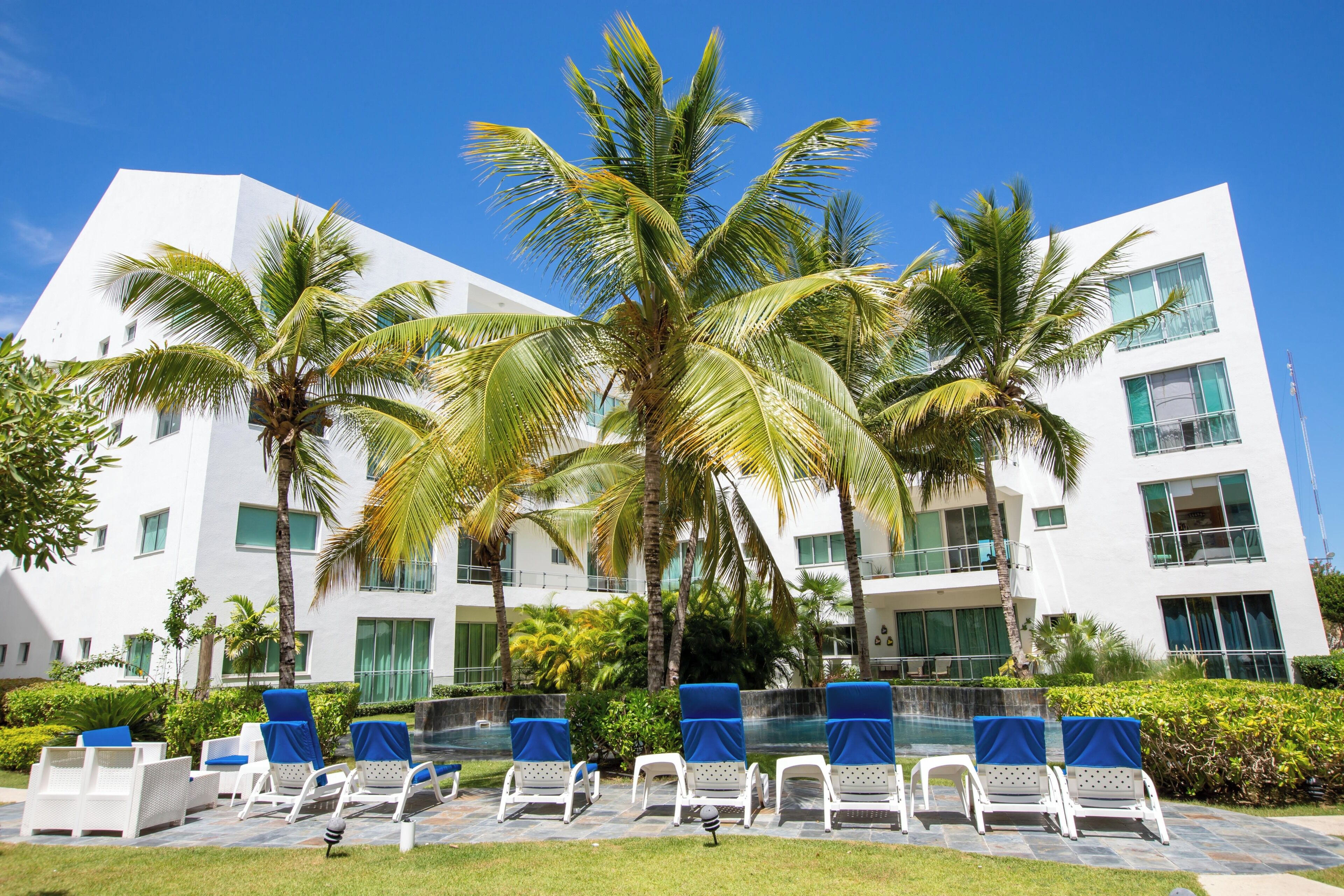 A modern building with multiple floors, a pool area with lounge chairs, and palm trees.