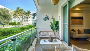 A balcony with a sofa, table, and decorative items, overlooking a lush garden and a building with palm trees. - King Size Bed| PrivateBBQ| CapCana| Beach Juanillo (Punta Cana)