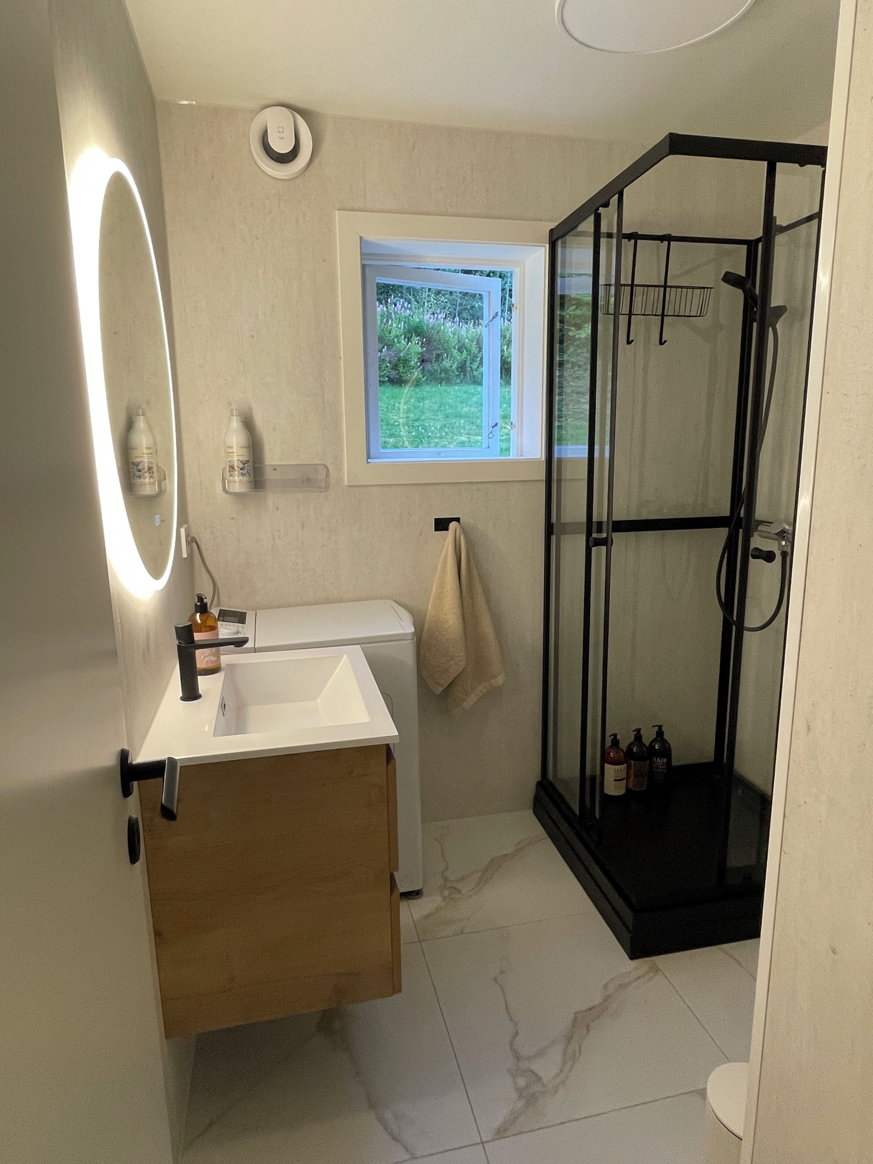 Ein modernes Badezimmer mit Duschkabine aus Glas, einem weißen Waschbecken und einem Fenster mit Blick auf Grünflächen.