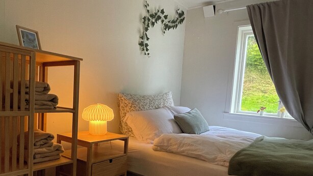 A bedroom with a bed, a bedside table with a lamp, a shelf with folded towels, and a window with curtains. - Solvang Ferienhaus mit Fjordblick & privatem Garten (Møre og Romsdal)