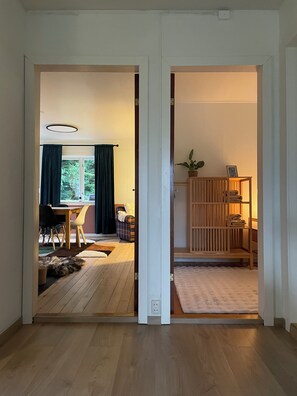 A hallway with a wooden floor leading to a dining area with a table and chairs, a sofa, and a window with curtains.