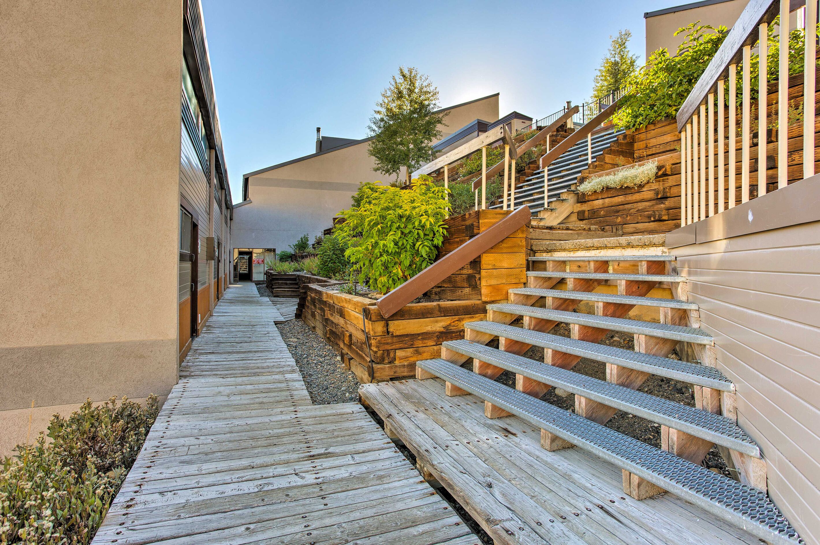 A narrow wooden pathway leading to a set of outdoor stairs with a wooden handrail and a metal safety handrail.