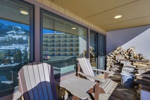 A balcony with two Adirondack chairs, a stack of firewood, and a view of a mountain resort.