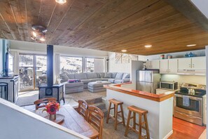 A spacious living area with a wooden ceiling, a kitchen with modern appliances, and a large window overlooking a snowy landscape.