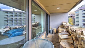 Ein Balkon mit Glastür, Tisch und Stuhl. Holzscheite und ein Metalleimer sind gestapelt.
