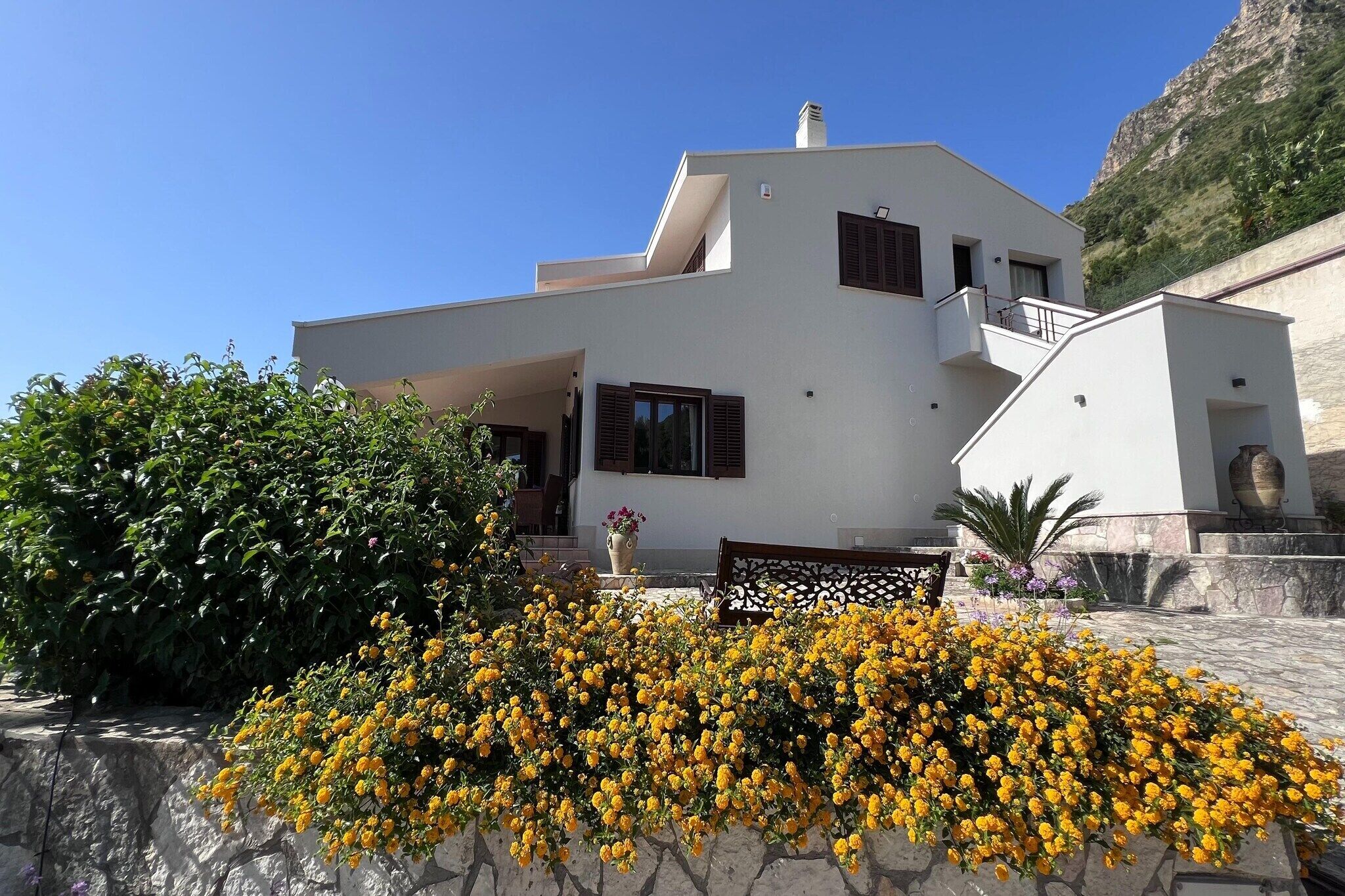 Une maison moderne à deux étages, dotée d’un balcon et entourée d’un jardin où poussent des fleurs jaunes.