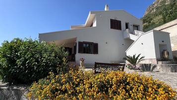 Une maison moderne à deux étages, dotée d’un balcon et entourée d’un jardin où poussent des fleurs jaunes.