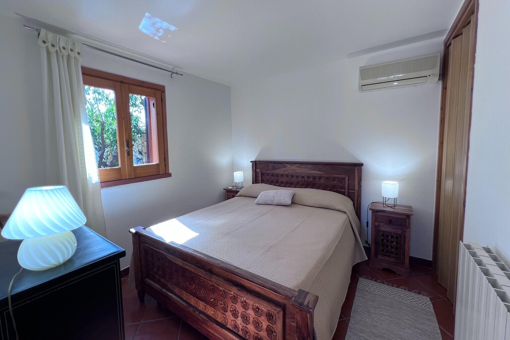 Een slaapkamer met een houten bed, een nachtkastje met lamp, een raam met gordijnen en een radiator.