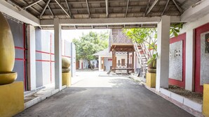 Uma galeria coberta com piso de concreto, que conduz a um edifício com fachada vermelha e branca, e uma estrutura de madeira com escadas.