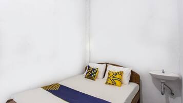 Quarto pequeno com uma cama de solteiro, cabeceira de madeira, paredes brancas, colcha branca, duas almofadas com padrões geométricos e pia branca com espelho.