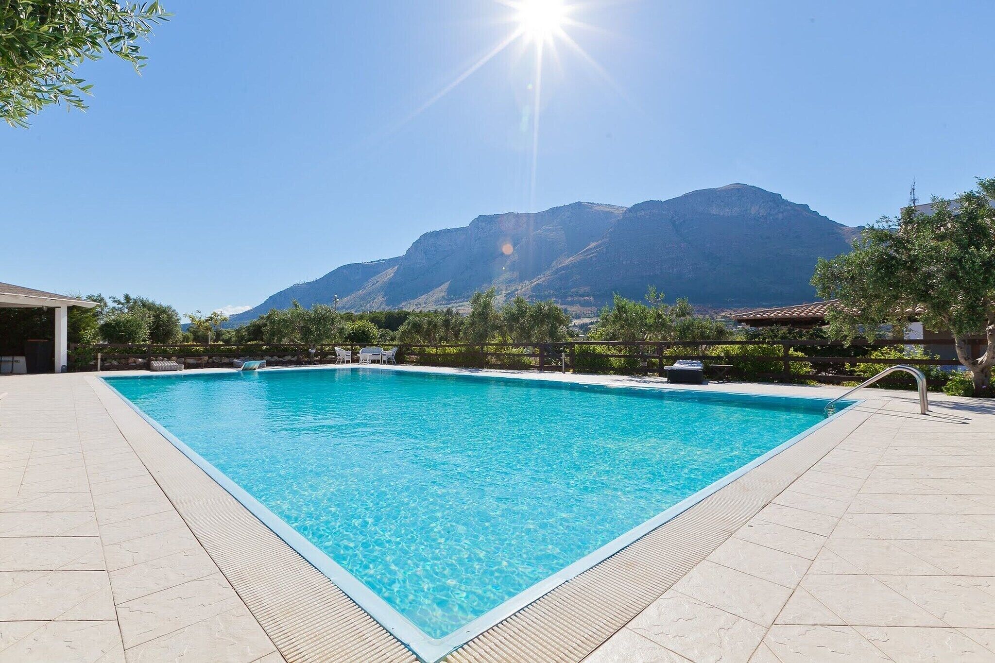 Una piscina rettangolare con acqua cristallina, circondata da un solarium piastrellato, con montagne e un cielo sereno sullo sfondo.