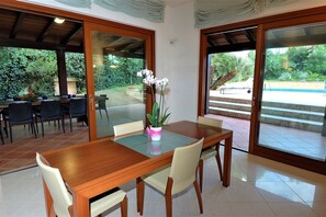 An indoor dining area with a wooden table and chairs, a potted orchid, and a view of a swimming pool through glass doors. - Villa in Valderice with Pool and Garden (Valderice (TP))