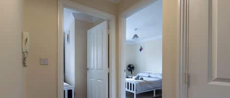 A hallway leading to a bedroom with a bed, a bedside table, a fan, and a wall-mounted telephone.