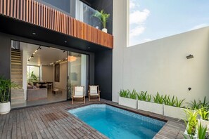 A modern outdoor pool area with a wooden deck, two lounge chairs, and a glass door leading to an interior space. - Cozy 2BR Villa in Umalas | Near Seminyak & Canggu (Umalas)