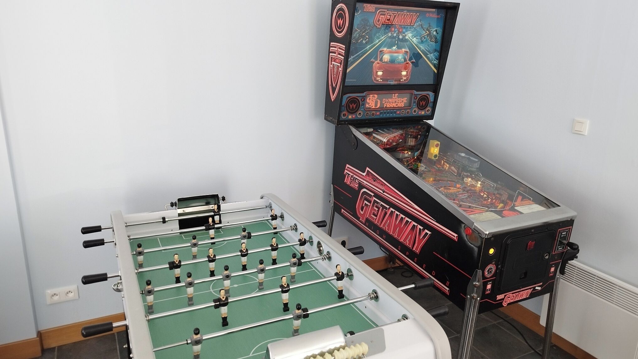 Una mesa de fútbol de mesa y una máquina de pinball en una habitación con paredes de colores claros.