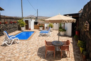 A pool area with lounge chairs and a table, surrounded by potted plants and a stone wall. - Holiday Home in Lajares for 5 (Lajares)