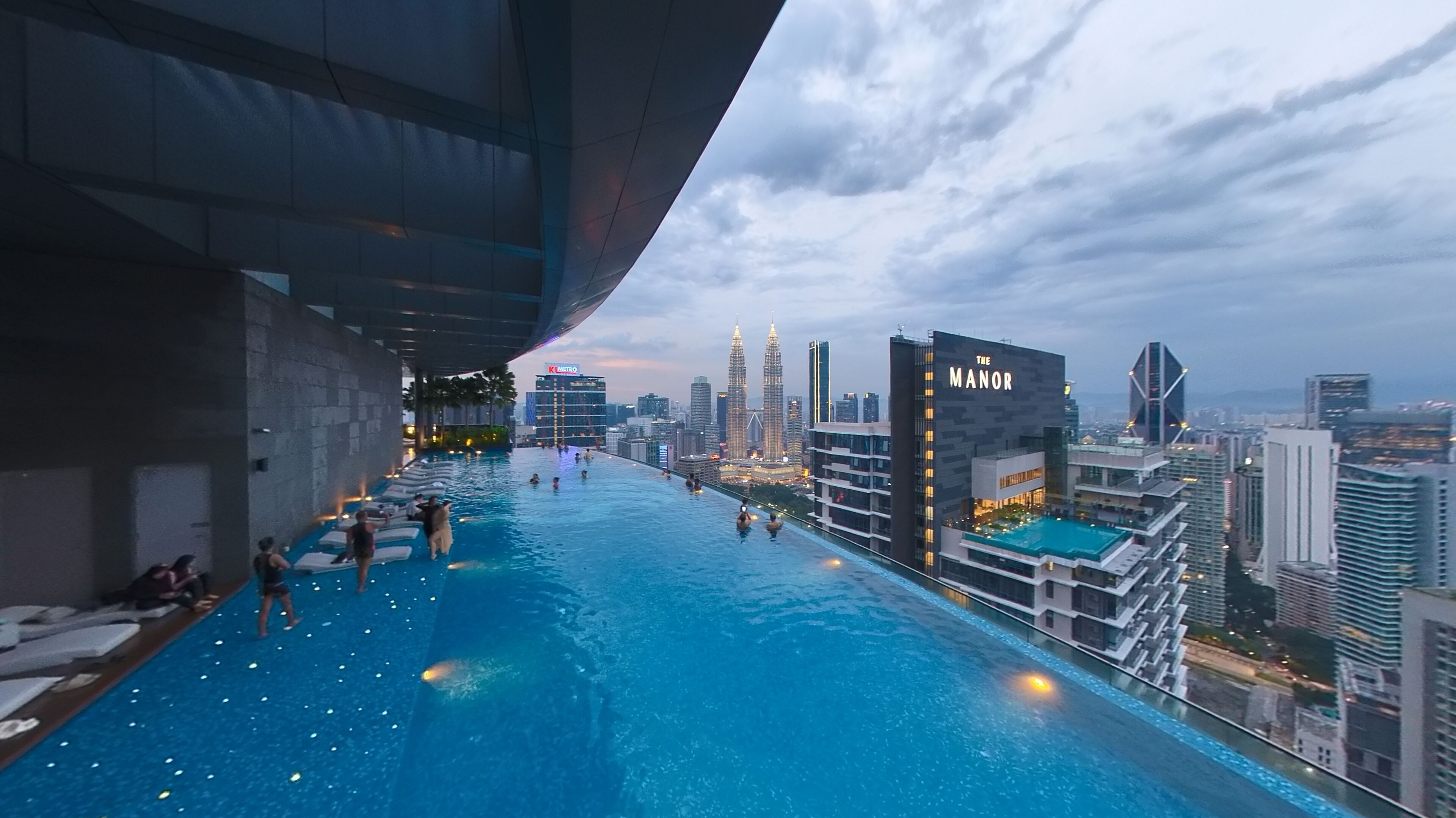 Ein Infinity-Pool mit Blick auf eine Stadtlandschaft, darunter die Petronas Towers, und ein Gebäude mit der Aufschrift 'The Manor'.