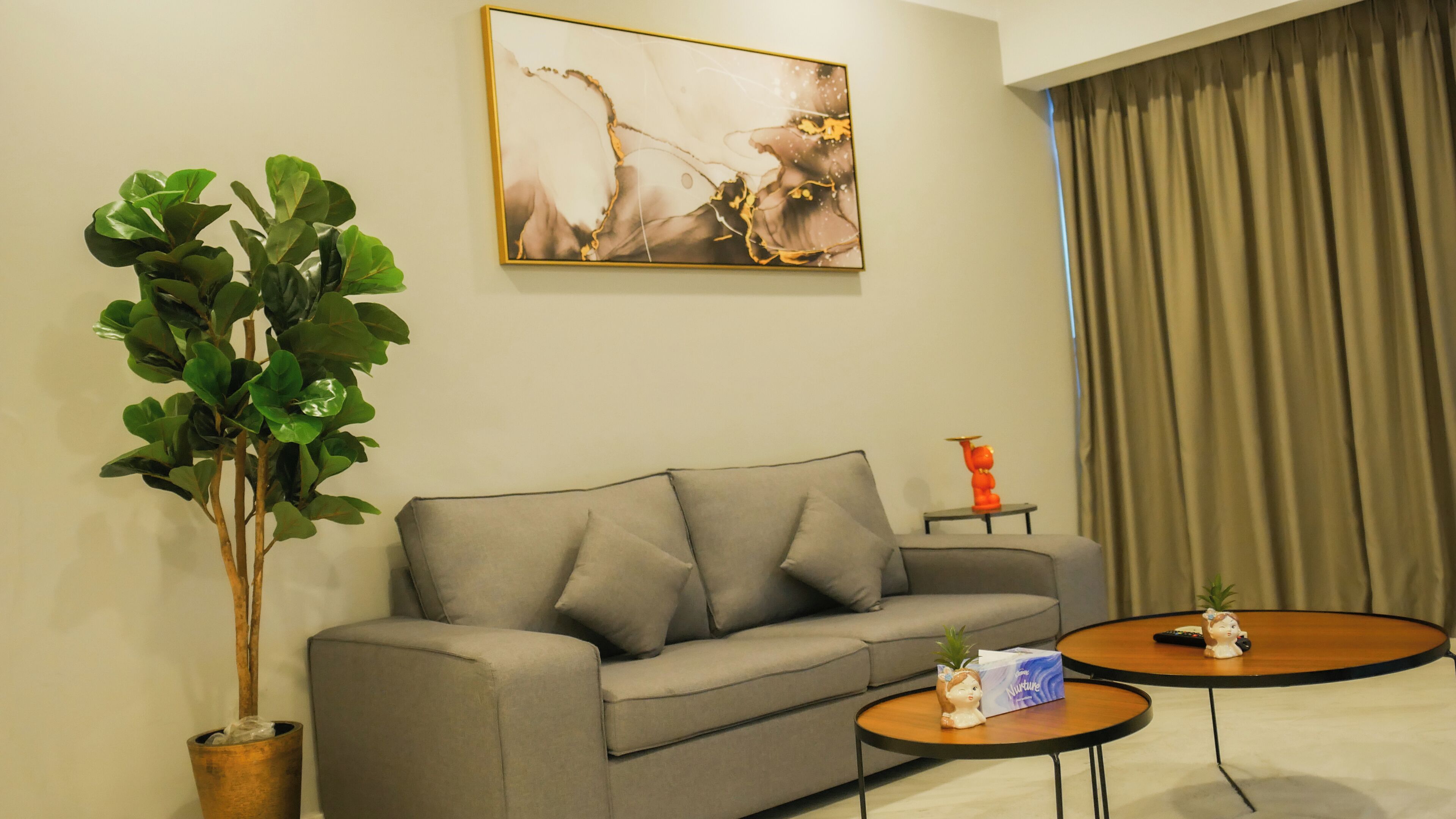 A living room with a grey sofa, a coffee table, a potted plant, a painting on the wall, and a curtain.