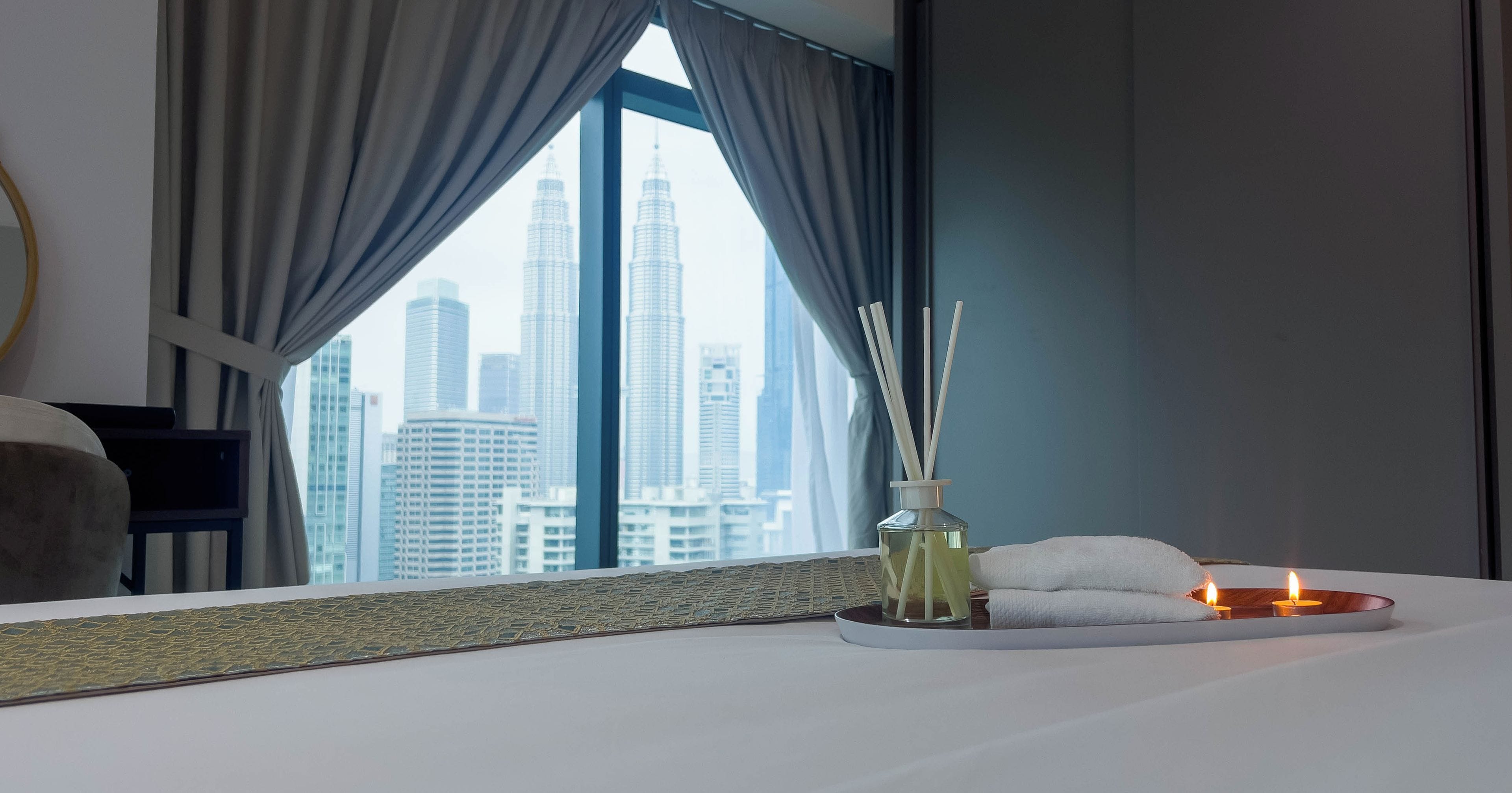 A hotel room with a large window, a bed with candles, a vase with reed sticks, and a towel.