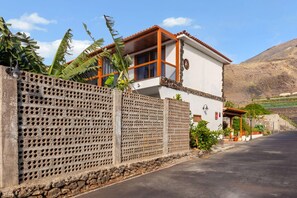 Une maison à deux étages avec un balcon, un mur de pierre orné d’un motif en treillis et une montagne en arrière-plan.