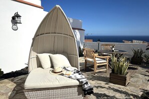 En mysig utomhusplats med sittmöjligheter, bestående av en vadderad bänk, en trästol och ett bord med en flaska på.